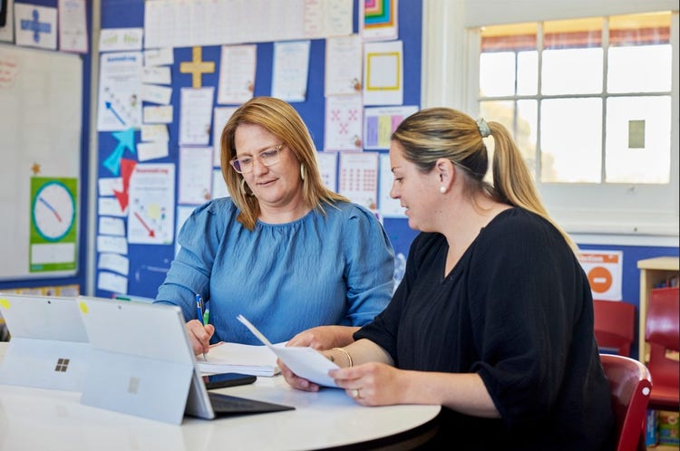 Two_female_educators_sit_at_a_desk_with_open_tablets_and_look_at_a_piece_of_paper